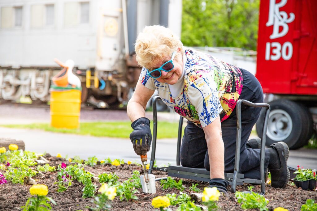 An older woman with blonde hair, wearing blue framed glasses and a multi-colored shirt, gardening outside.