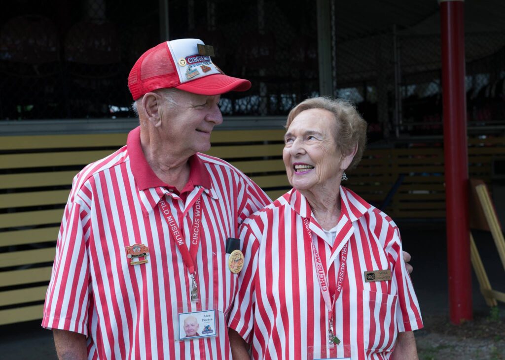 An older man, smiling and outside, wearing a red and white "Circus World" hat and a red and white striped shirt looking at and has his arm around an older woman with light brown hair and wearing a red and white striped shirt.