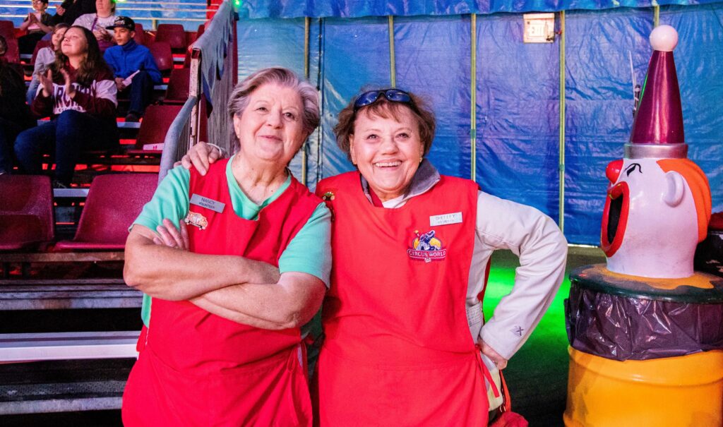 Two older women smiling and wearing a red "Circus World" apron, with the circus bleachers in the background.