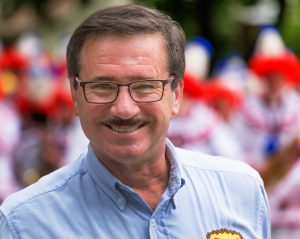 A headshot of a smiling man with brown hair and a mustache, wearing thin dark framed glasses and a blue collared shirt, with people blurred out in the background.